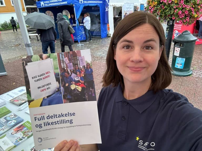 Bianca Løkkeberg har halvlangt brunt hår. På bildet smiler hun og holder opp et SAFOmagasin. Bildet er tatt på Arendalsuka i 2023.